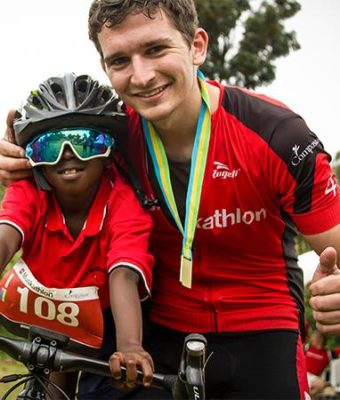 Deelnemer van de Muskathlon poseert met Afrikaans jongetje die met zijn zonnebril op op zijn mountainbike zit