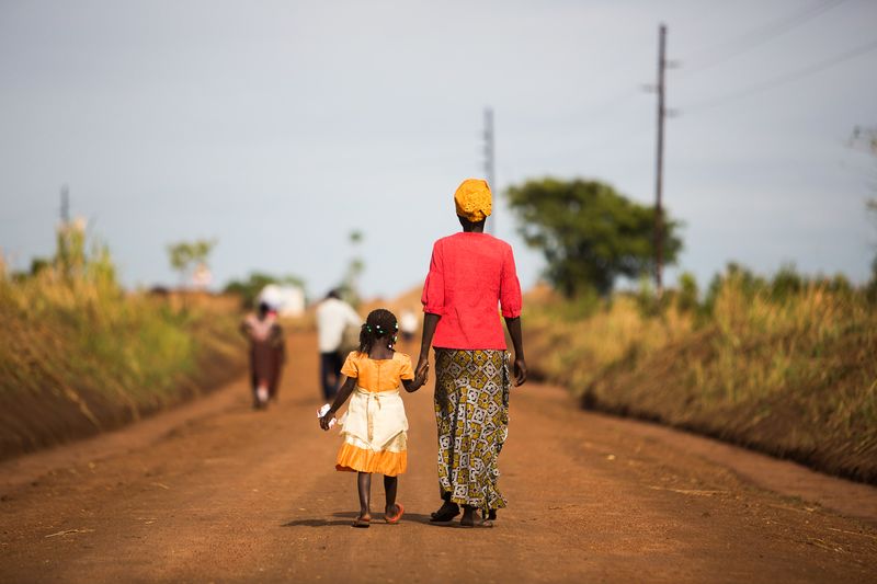 Vrouw met oranje hoofddoek, rode bloes en wit/gele rok loopt hand in hand met meisje in oranje jurk over rode zandweg in Uganda