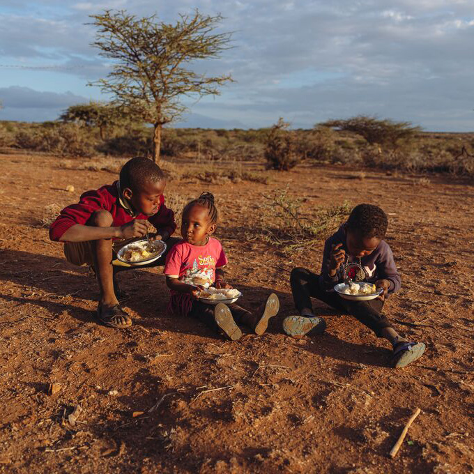 Drie kinderen zitten met een bord eten in het zand.