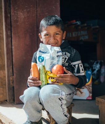 Een jongen houdt een voedselpakket vast en zit op de drempel van zijn huis.
