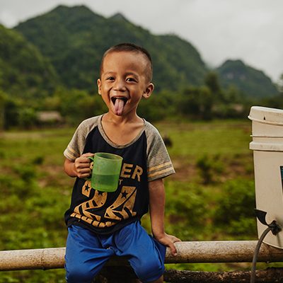 Een jongetje uit Thailand zit op een boomstam naast een waterfilter en houdt een beker water vast.