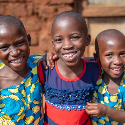 De Rwandese pleegzussen Gisele, Impano en Umutesiwase staan gearmd en lachen naar de camera.