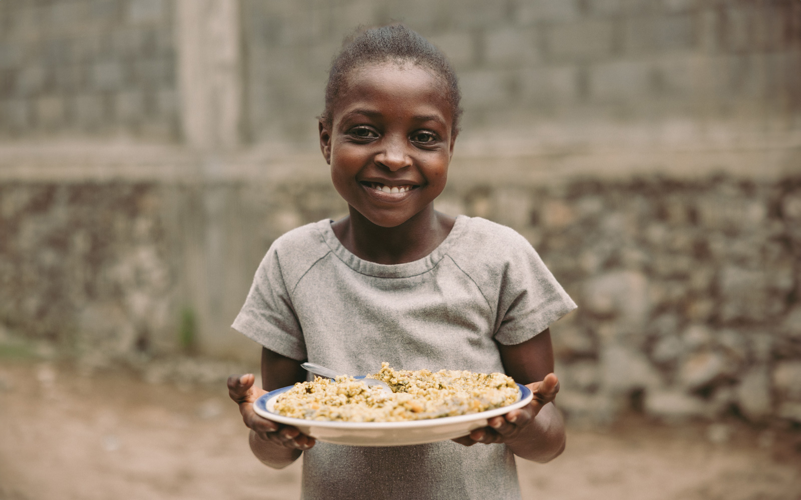 Rose Mirline uit Haïti staat voor een stenen muur en houdt een bord eten vast.