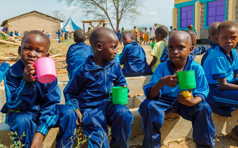 Een groep Keniaanse kinderen zit op een muurtje en drinkt uit bekers.