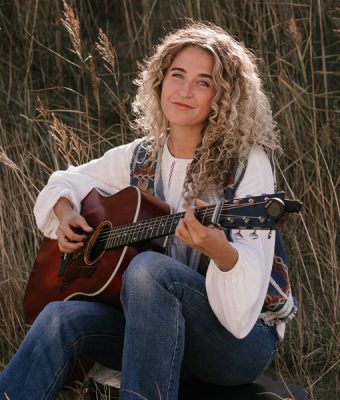Singer-songwriter Sera Noah zit in het gras en speelt op haar gitaar.