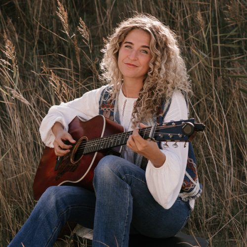 Singer-songwriter Sera Noah zit in het gras en speelt op haar gitaar.