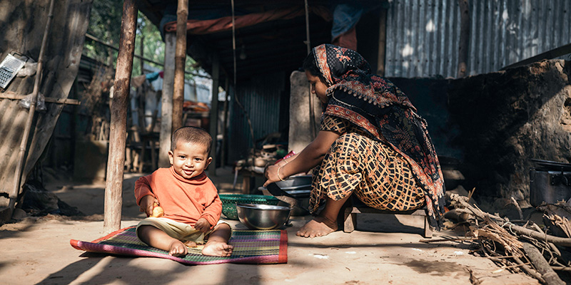 Een vrouw uit Bangladesh bereidt eten voor haar huis terwijl haar zoon op een kleed zit.