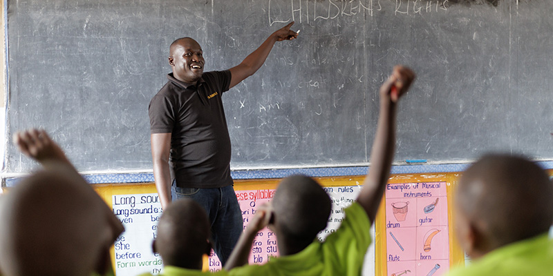 Projectdirecteur Moses uit Uganda wijst iets aan op een schoolbord in een klaslokaal.